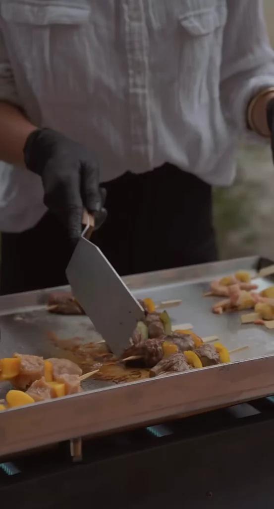 Brochettes de viande et de légumes cuisinées sur une plancha, manipulées à la spatule par un cuisinier.
